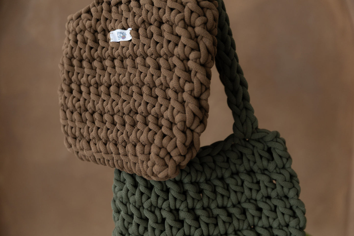 2 gehäkelte mittelgroße Schultertaschen in den Farben Nussbraun und Avokadogrün hängd und stark vergrößert, so dass nicht die kom pletten Taschen zu sehen sind. Im Hintergrund eine braune Limewash-Wand.