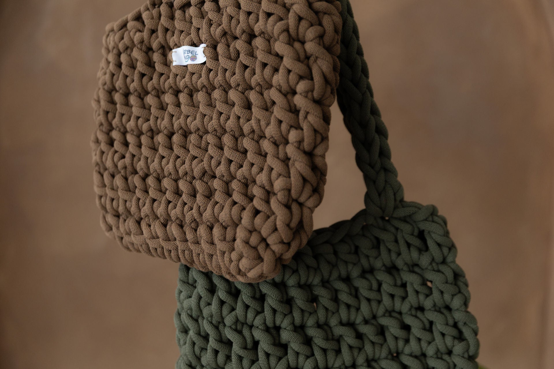 2 gehäkelte mittelgroße Schultertaschen in den Farben Nussbraun und Avokadogrün hängd und stark vergrößert, so dass nicht die kom pletten Taschen zu sehen sind. Im Hintergrund eine braune Limewash-Wand.