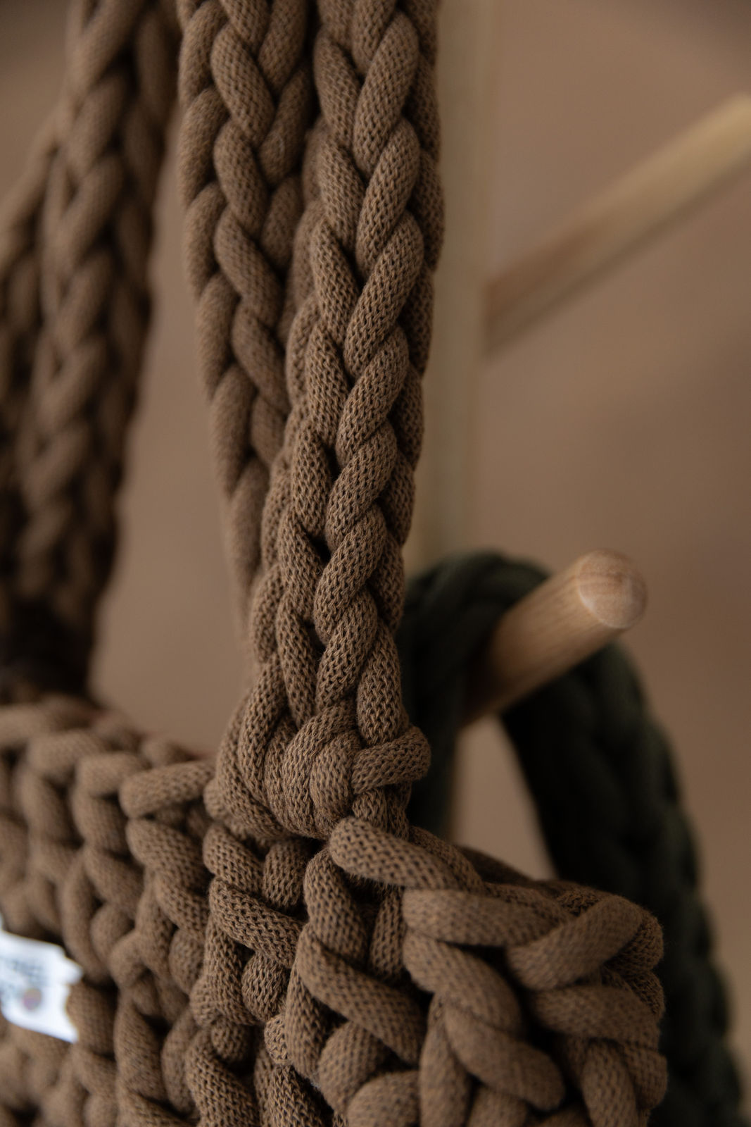 2 gehäkelte mittelgroße Schultertaschen in den Farben Nussbraun und Avokadogrün hängen an einem Garderobenständer aus Holz. Im Hintergrund eine braune Limewash-Wand mit einer Bogen-Nische. Die Taschen sind sehr stark vergrößert, wir sehen fast ausschließlich die Schulterriemen der braunen und das sehr dicke Garn aus dem sie gemacht sind. Der Hintergrund ist verschwommen.