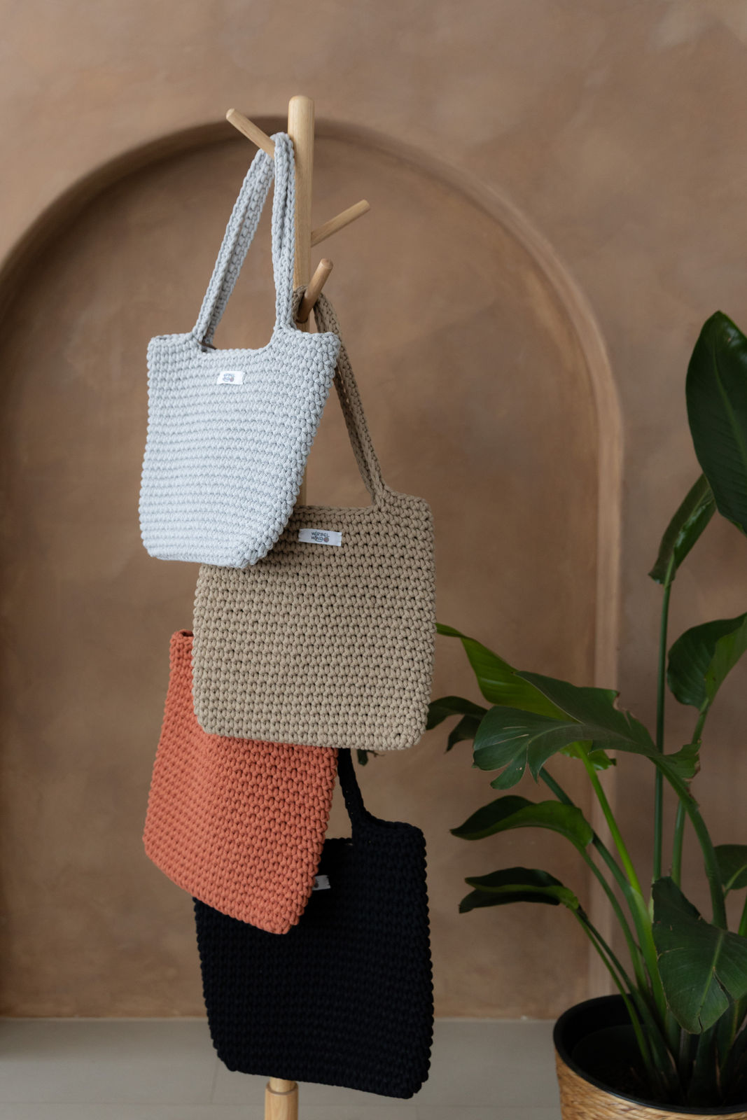 4 gehäkelte große Schultertaschen in den Farben Schwarz, Grau, Beige und Terracotta Orange hängen an einem Garderobenständer aus Holz. Im Hintergrund eine braune Limewash-Wand mit einer Bogen-Nische. Auf dem Boden am rechten Rand steht eine Pflanze mit großen grünen Blättern in einem hellbraunen Topf, das Material ähnlich Bambus.