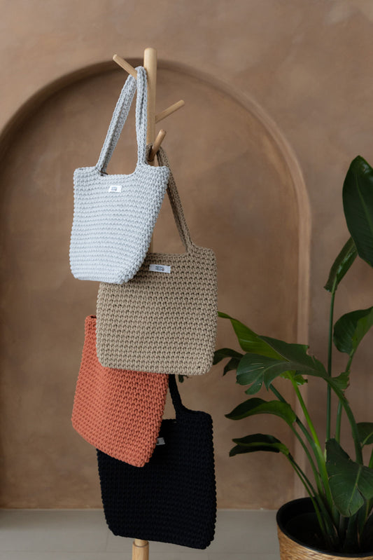 4 gehäkelte große Schultertaschen in den Farben Schwarz, Grau, Beige und Terracotta Orange hängen an einem Garderobenständer aus Holz. Im Hintergrund eine braune Limewash-Wand mit einer Bogen-Nische. Auf dem Boden am rechten Rand steht eine Pflanze mit großen grünen Blättern in einem hellbraunen Topf, das Material ähnlich Bambus.