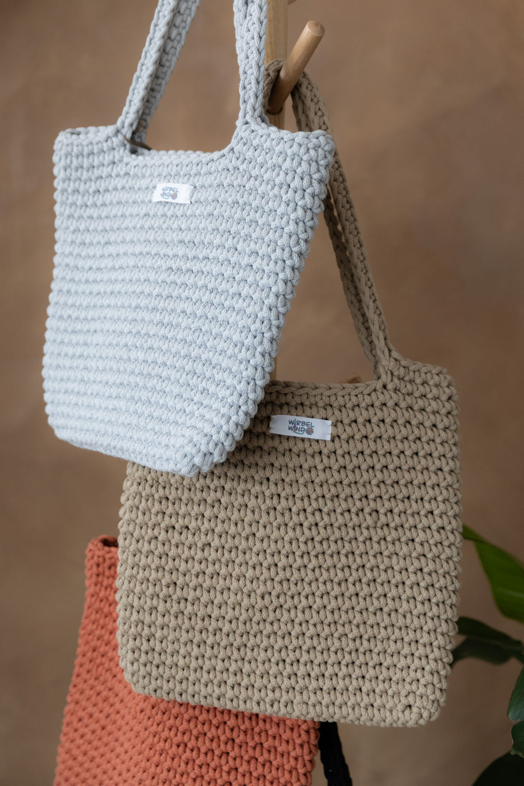 4 gehäkelte große Schultertaschen in den Farben Schwarz, Grau, Beige und Terracotta Orange hängen an einem Garderobenständer aus Holz. Im Hintergrund eine braune Limewash-Wand mit einer Bogen-Nische. Auf dem Boden am rechten Rand steht eine Pflanze mit großen grünen Blättern in einem hellbraunen Topf, das Material ähnlich Bambus. Gleich dem vorherigen Bild, nur eine viel nähere Aufnahme, so dass nur ein Ausschnitt und nicht alle Taschen sichtbar sind.