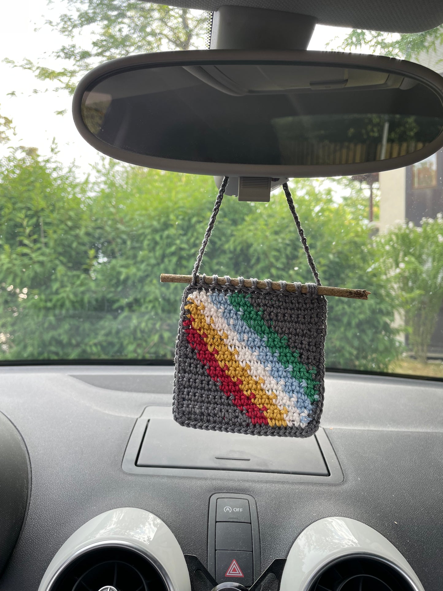 Die gehäkelte Flagge hängt in einem Auto an einem Rückspiegel. Wir sehen das Armaturenbrett, den Spiegel, im Hintergrund (vor Auto) verschwommen ein grüner Busch.