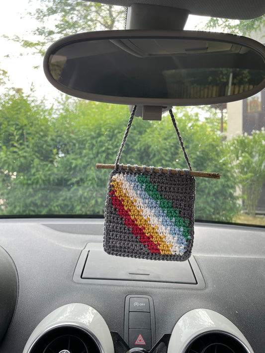Die gehäkelte Flagge hängt in einem Auto an einem Rückspiegel. Wir sehen das Armaturenbrett, den Spiegel, im Hintergrund (vor Auto) verschwommen ein grüner Busch.