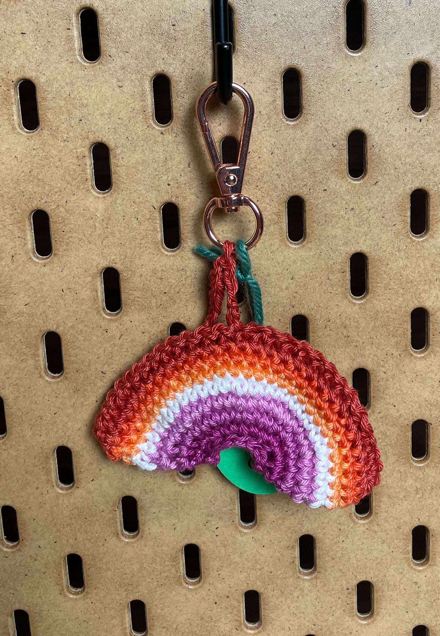 Gehäkelter Regenbogen Schlüsselanhänger mit Karabiner in den 7 Farben der Lesbischen Pride Flagge: Orangerot, Orange, Hellorange, Weiß, Hellpink, Mitelpink, Dunkelpink. Außerdem hängt das Logo von Wirbelwind ebenfalls am Karabiner.