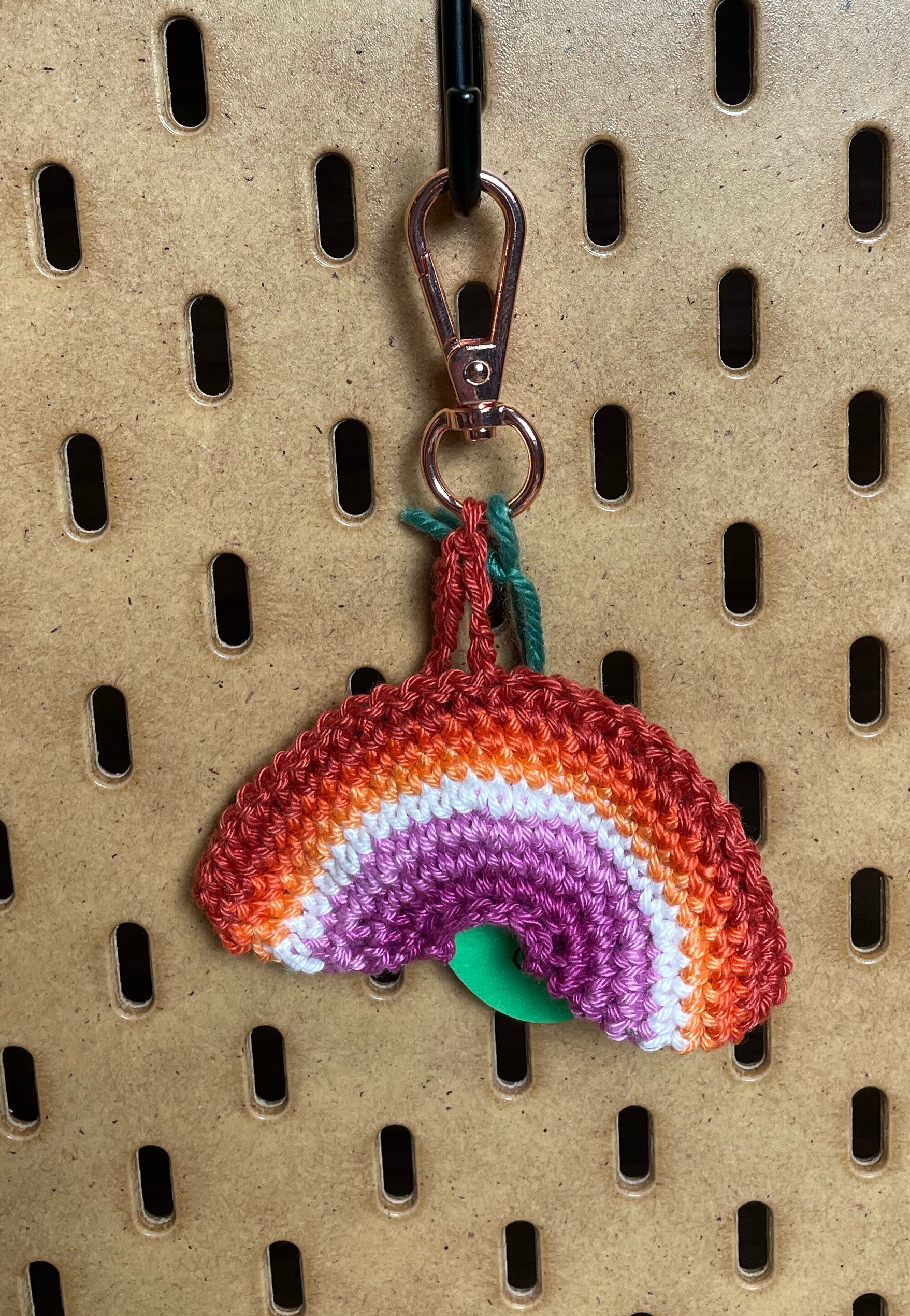 Gehäkelter Regenbogen Schlüsselanhänger mit Karabiner in den 7 Farben der Lesbischen Pride Flagge: Orangerot, Orange, Hellorange, Weiß, Hellpink, Mitelpink, Dunkelpink. Außerdem hängt das Logo von Wirbelwind ebenfalls am Karabiner.