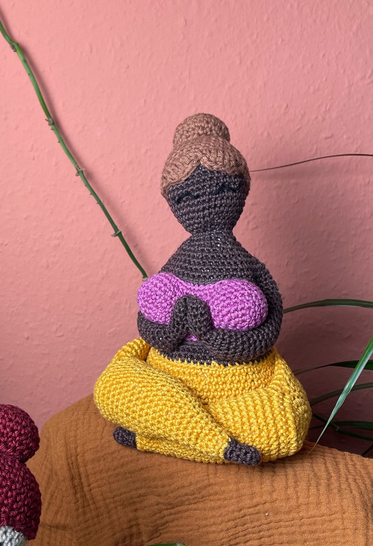 Gehäkelte Yoga Figur: hellbrauner Dutt, dunkelbraune Haut, pinkes Top, gelbe Hose