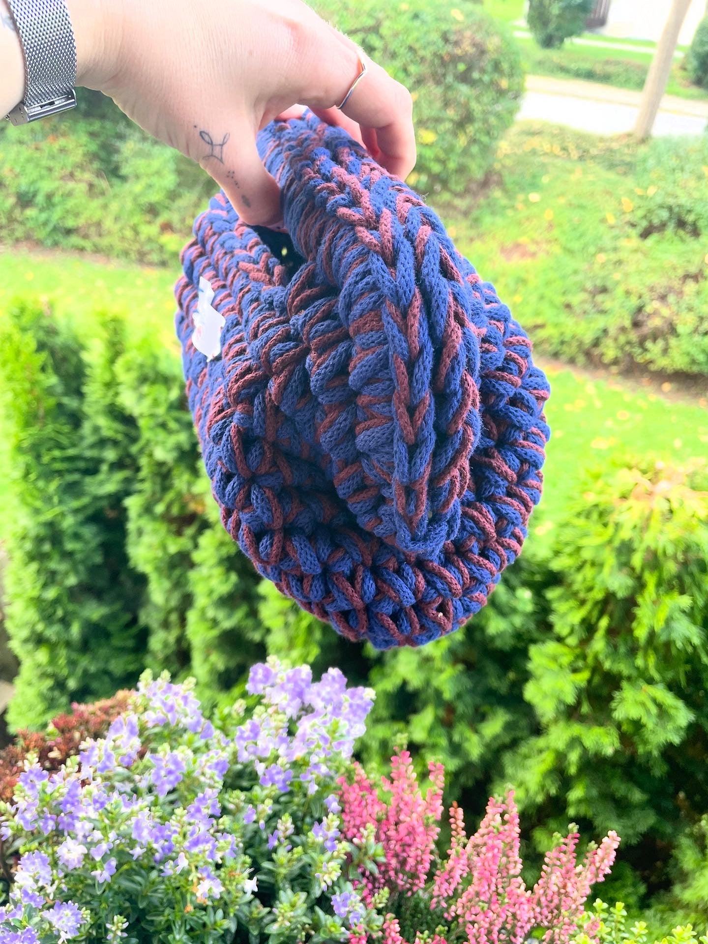 Aus dem linken Bildrand ragt ein Arm, der die Tasche am Griff über die Brüstung eines Balkons hält. Wir sehen die Tasche nun von der Seite und daher, wie bauchig sie ist. Im Hintergrund eine sonnenbeschienene Wiese und Blumen.
