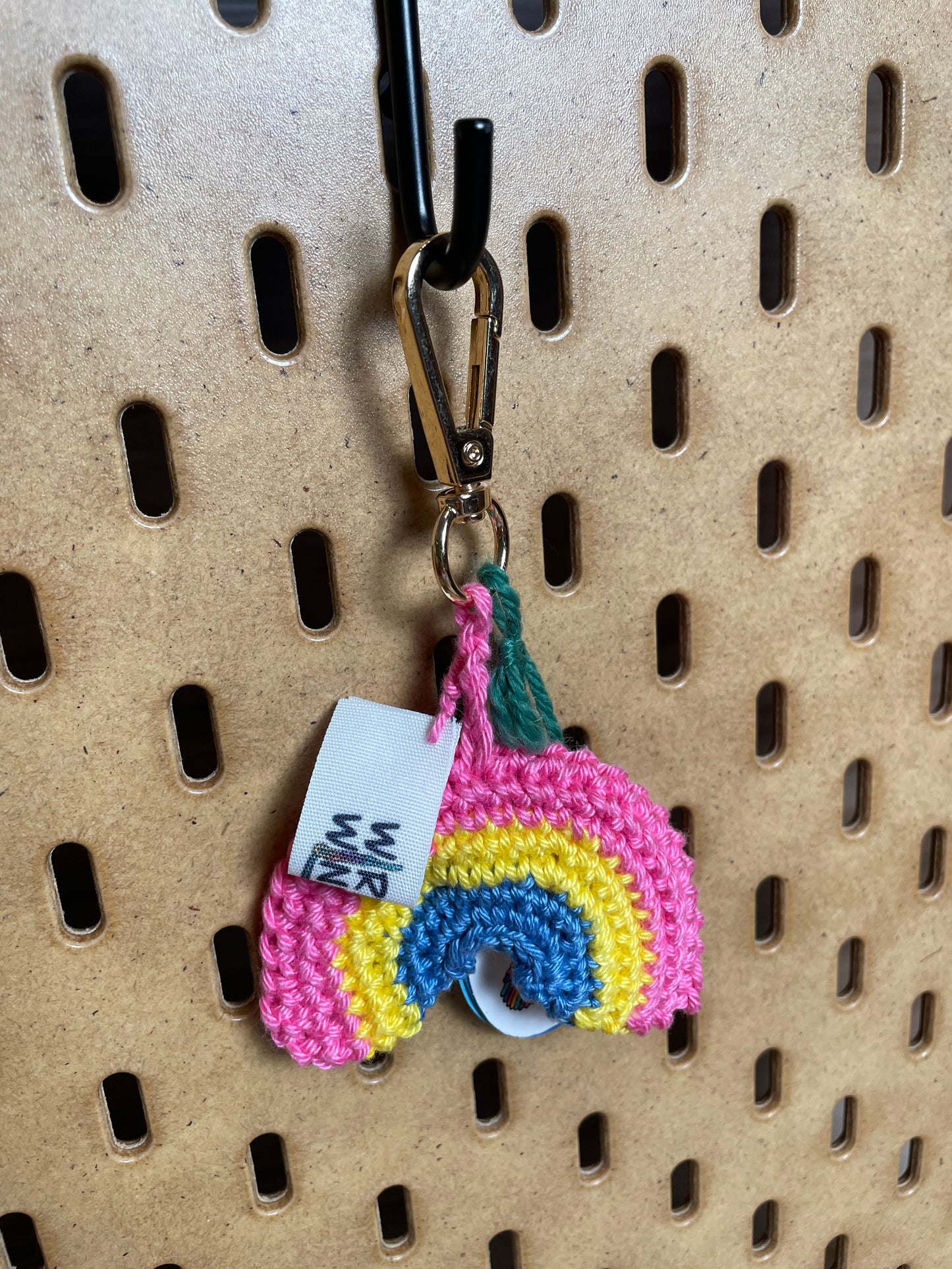 Gehäkelter Regenbogen Schlüsselanhänger mit Karabiner in den Farben der Pansexuellen Pride Flagge: Pink, Gelb, Blau. Außerdem hängt das Logo von Wirbelwind ebenfalls am Karabiner.