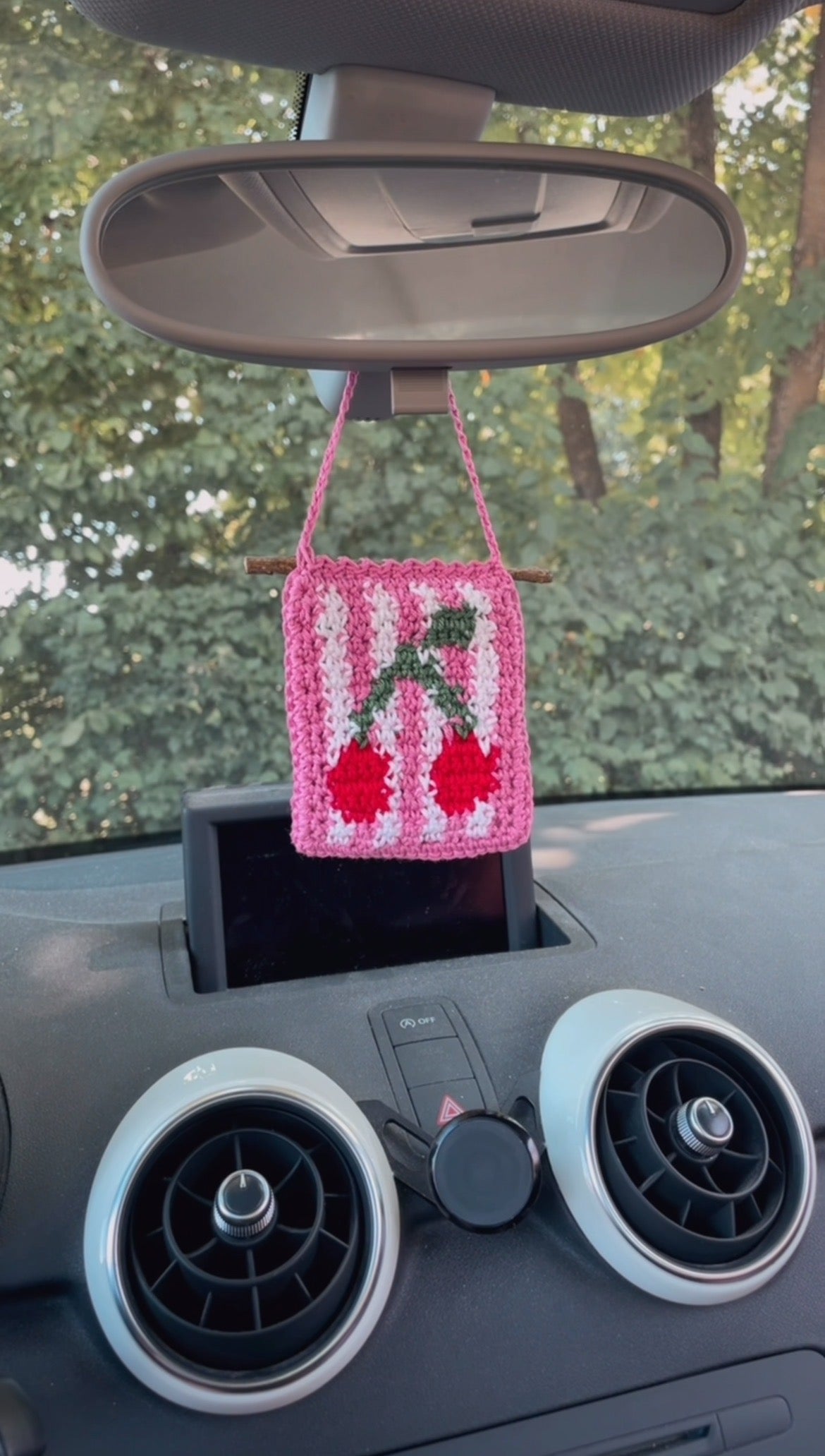 Gehäkelter Kirsch-Anhänger hängt in einem Auto an einem Rückspiegel: Im Vordergrund Kirsche, Hintergrund Weiß und Pink gestreift, umrandet von Pink, Aufhängung ebenfalls Pink.