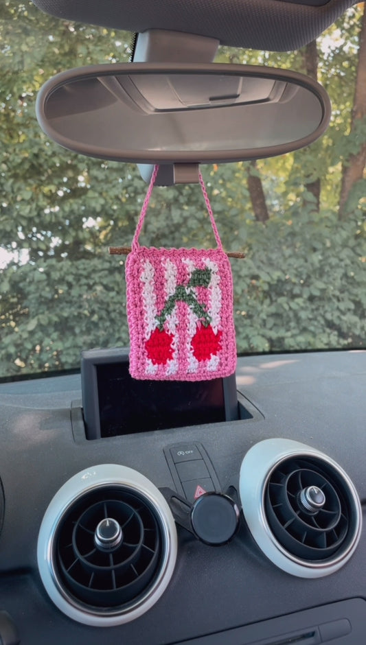 Gehäkelter Kirsch-Anhänger hängt in einem Auto an einem Rückspiegel: Im Vordergrund Kirsche, Hintergrund Weiß und Pink gestreift, umrandet von Pink, Aufhängung ebenfalls Pink.