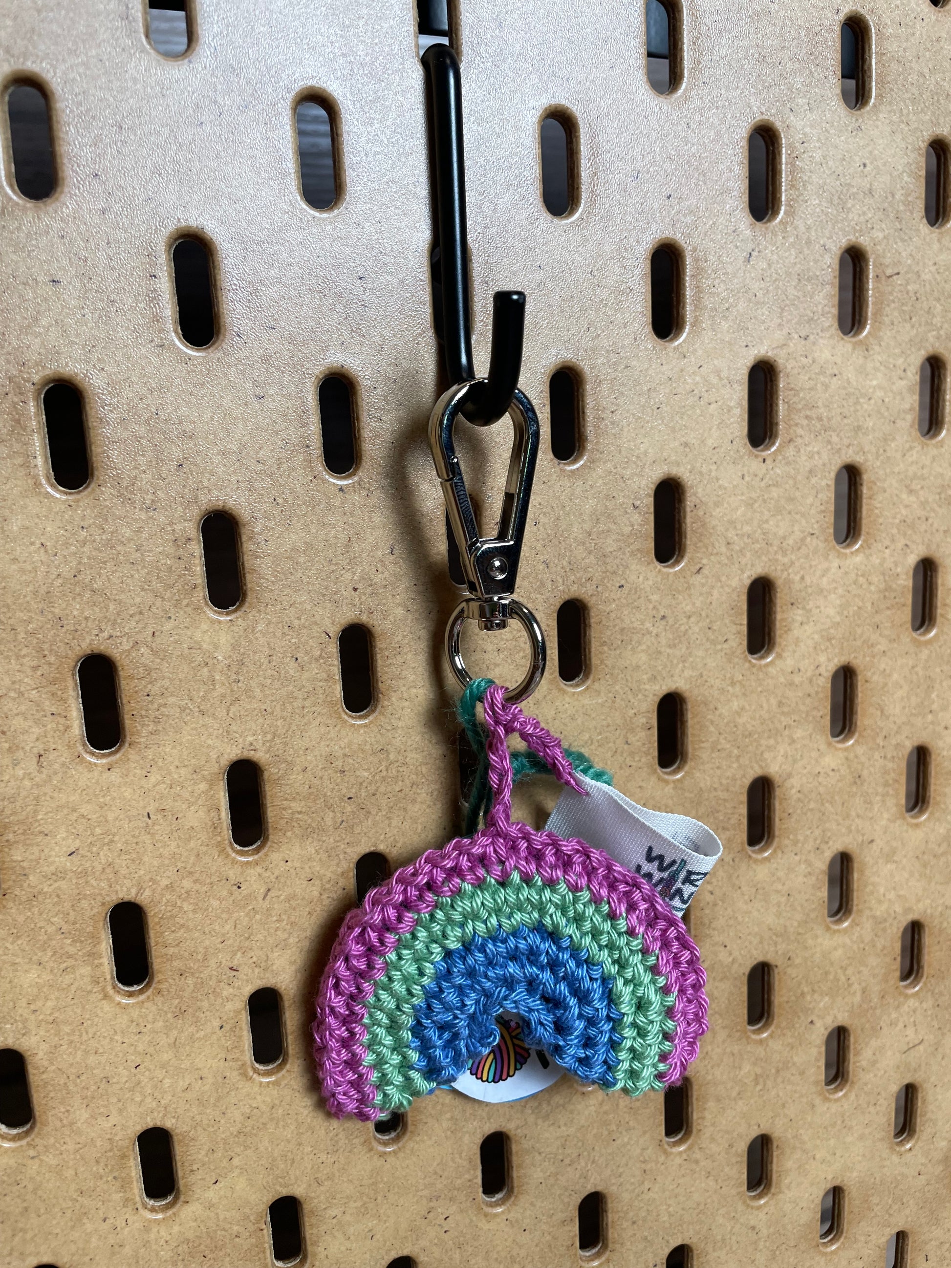 Gehäkelter Regenbogen Schlüsselanhänger mit Karabiner in den Farben der Polysexuellen Pride Flagge: Pink, Grün, Blau. Außerdem hängt das Logo von Wirbelwind ebenfalls am Karabiner.