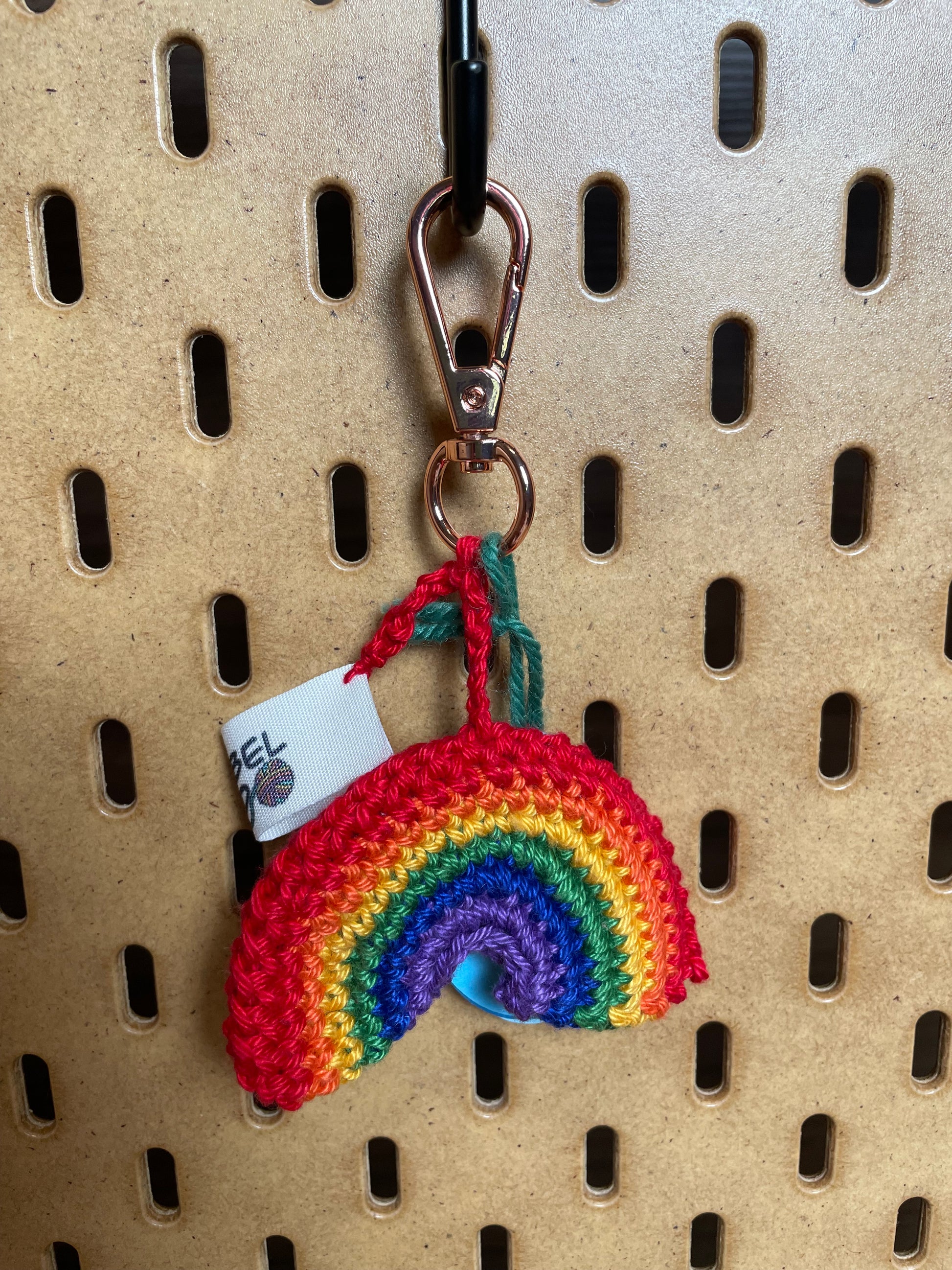 Gehäkelter Regenbogen Schlüsselanhänger mit Karabiner in den 6 Farben des Regenbogens: Rot, Orange, Gelb, Grün, Blau, etwas dunkleres Lila. Außerdem hängt das Logo von Wirbelwind ebenfalls am Karabiner.