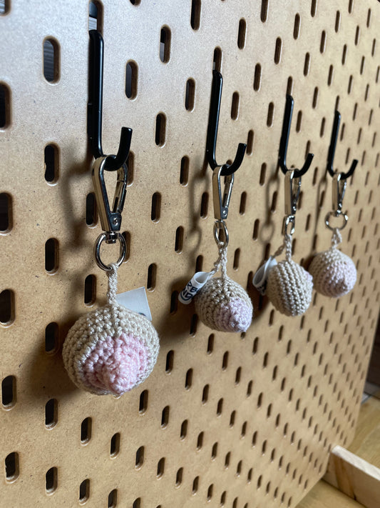 Seitliche Aufnahme: 4 gehäkelte Brüste in Hellbeige mit Rosa Nippeln/Narben an silbernen Karabinern aufgehängt.