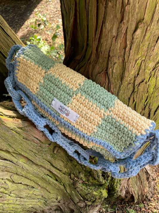 Die gelb-grün-karierte Tasche steht im Geäst eines Baumes, der blaue Kettengliedergriff hängt vorne nach unten und liegt auf dem Baum auf.