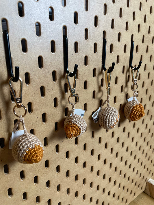 4 gehäkelte Brüste in Dunkelbeige mit orange-braunen Nippeln/Narben an goldenen Karabinern aufgehängt.