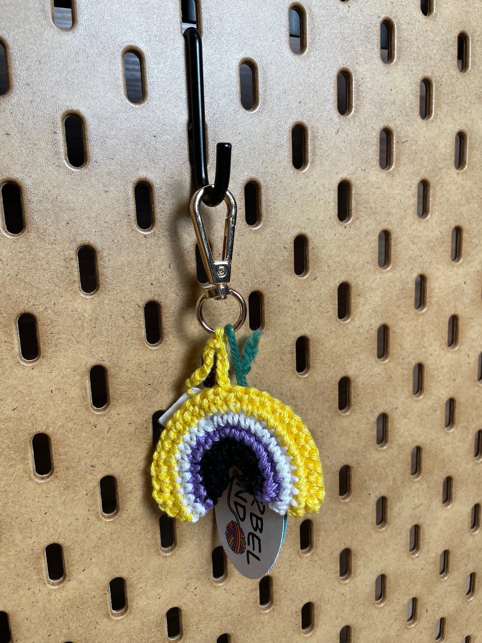 Gehäkelter Regenbogen Schlüsselanhänger mit Karabiner in den Farben der Nonbinary Pride Flagge: Gelb, Weiß, Lila, Schwarz. Außerdem hängt das Logo von Wirbelwind ebenfalls am Karabiner.