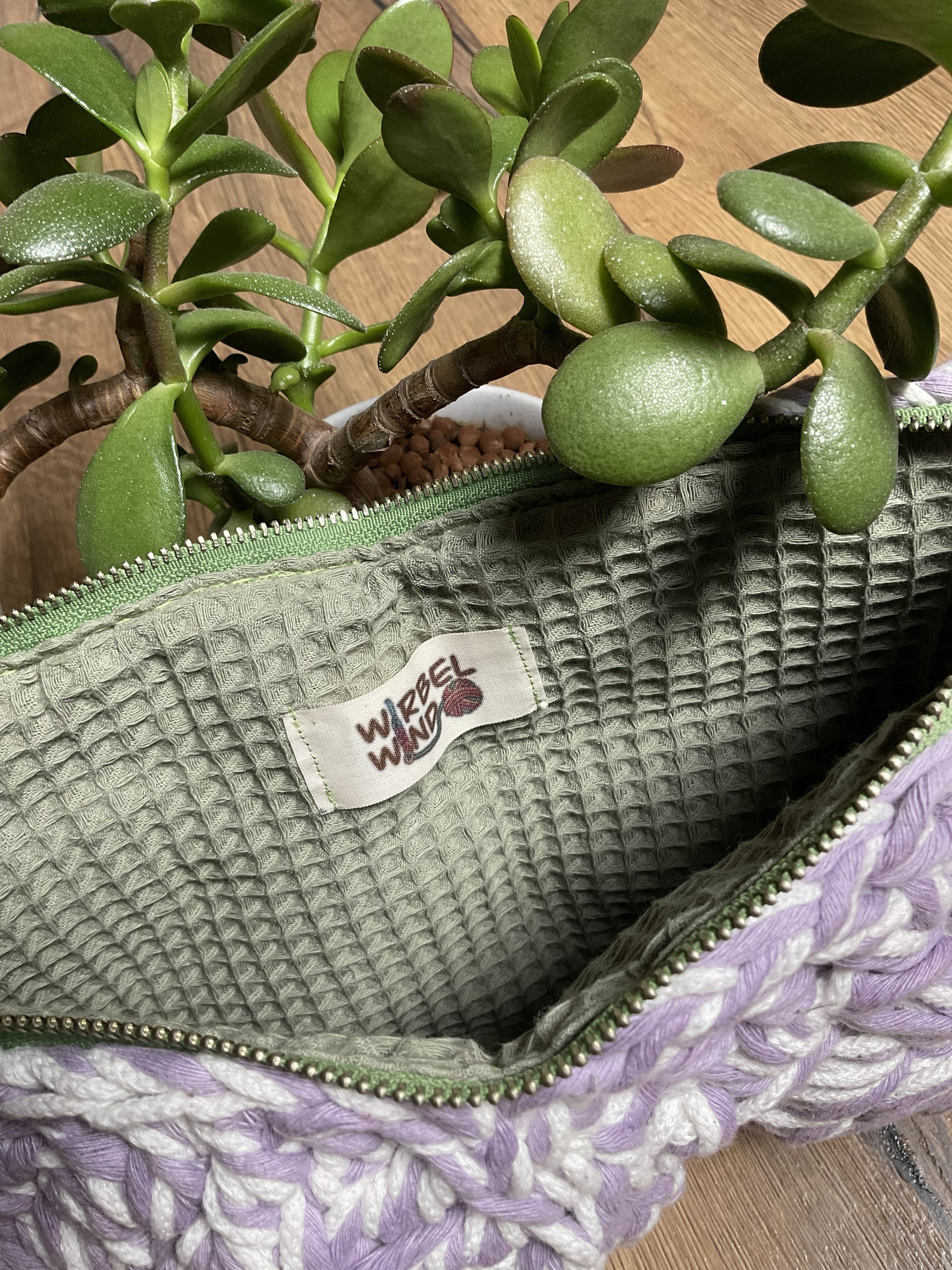 Der Reißverschluss der Tasche ist geöffnet, wir sehen das salbeigrüne Innenfutter aus Waffelstoff. Die Tasche steht neben einer Sukkulente.