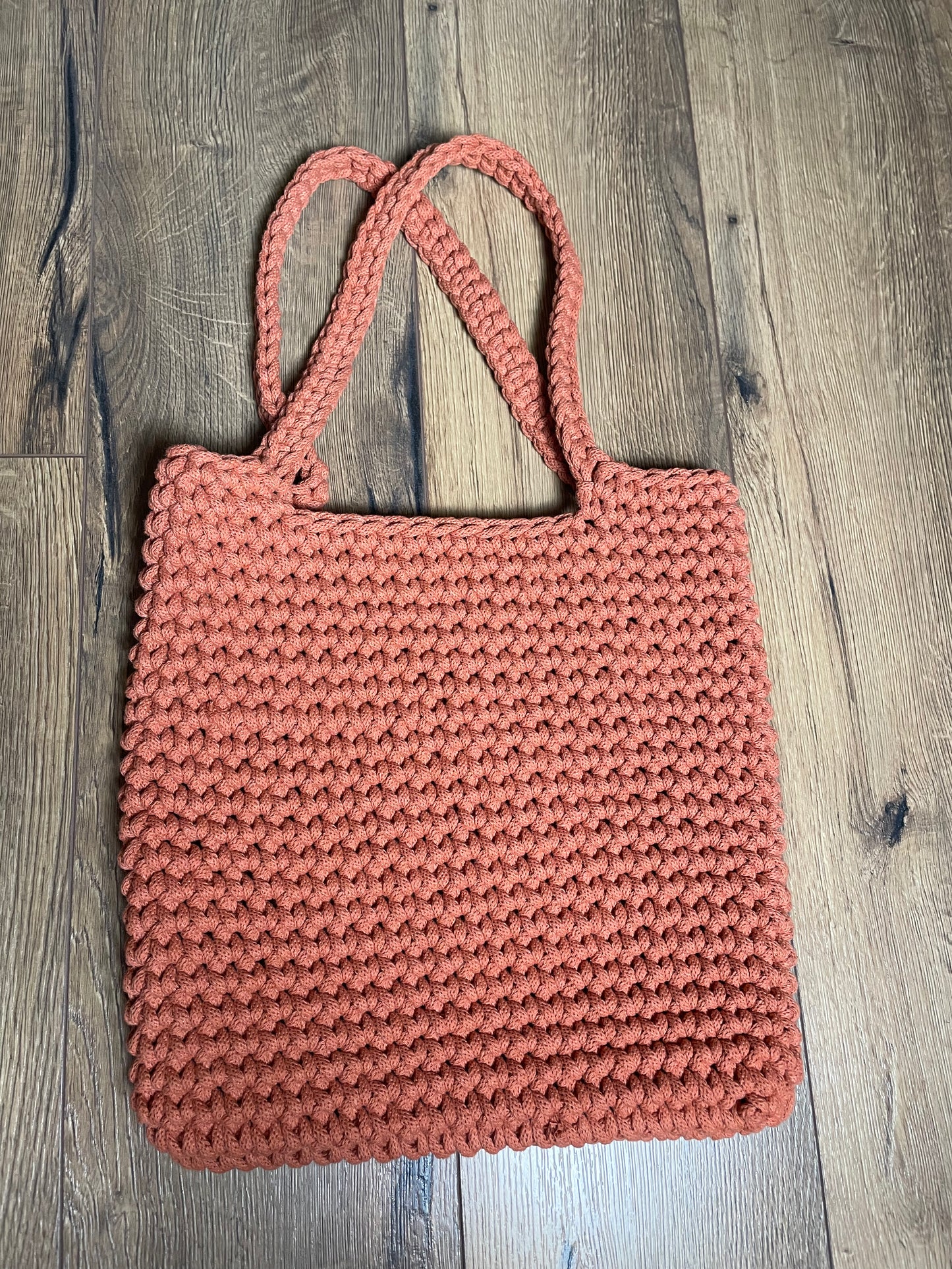 Eine terracotta-orangefarbene gehäkelte Schultertasche liegt auf Holzlaminatboden. Die Tasche hat ein gleichmäßiges Häkelmuster, zwei lange Henkel und eine rechteckige Form. Das handgefertigte Stück aus recyceltem Baumwollgarn zeigt die charakteristische Struktur der Häkelarbeit mit sichtbaren Maschen.
