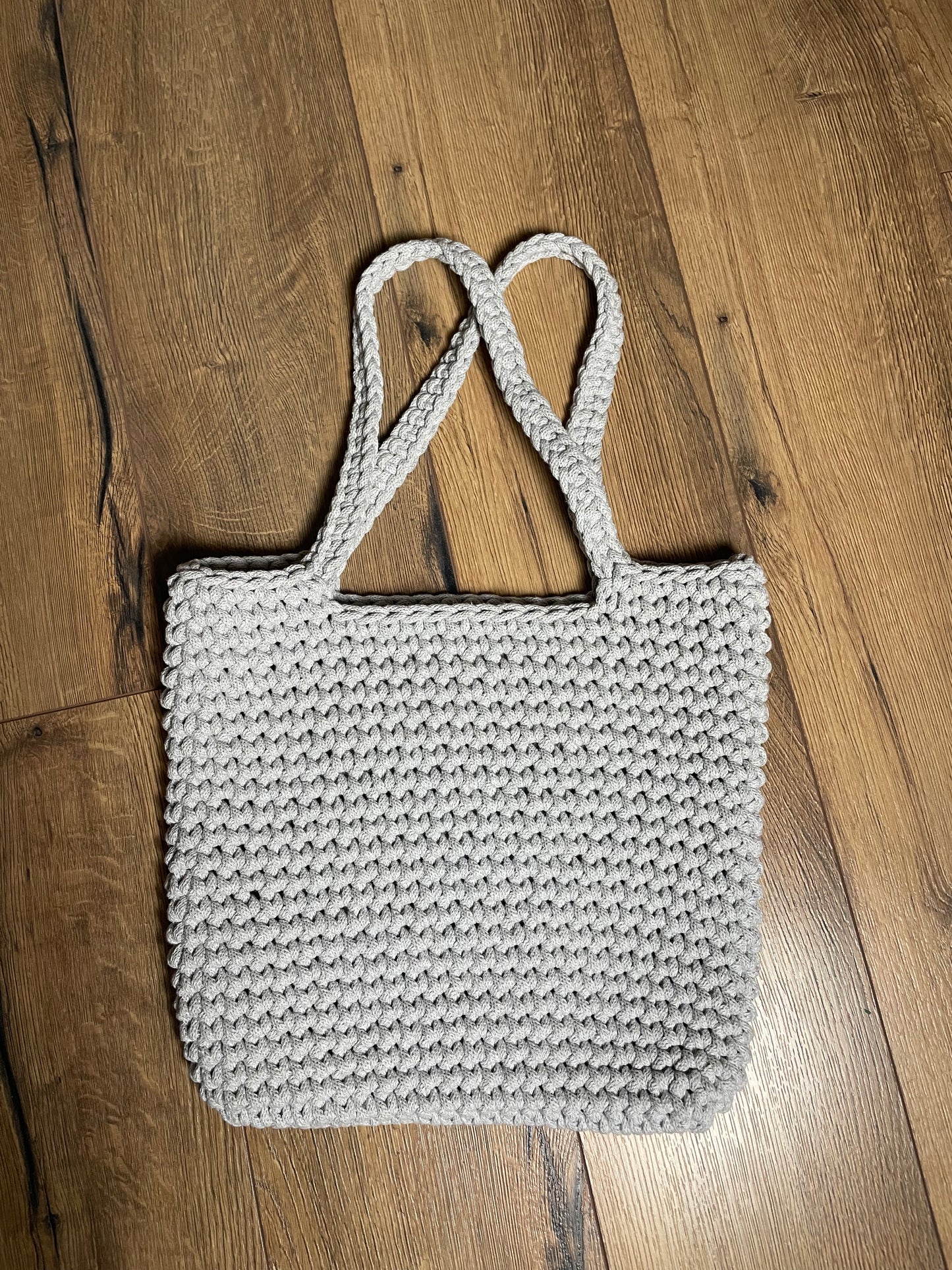 Eine hellgraue gehäkelte Schultertasche liegt auf Holzlaminatboden. Die Tasche hat ein gleichmäßiges Häkelmuster, zwei lange Henkel und eine rechteckige Form. Das handgefertigte Stück aus recyceltem Baumwollgarn zeigt die charakteristische Struktur der Häkelarbeit mit sichtbaren Maschen.
