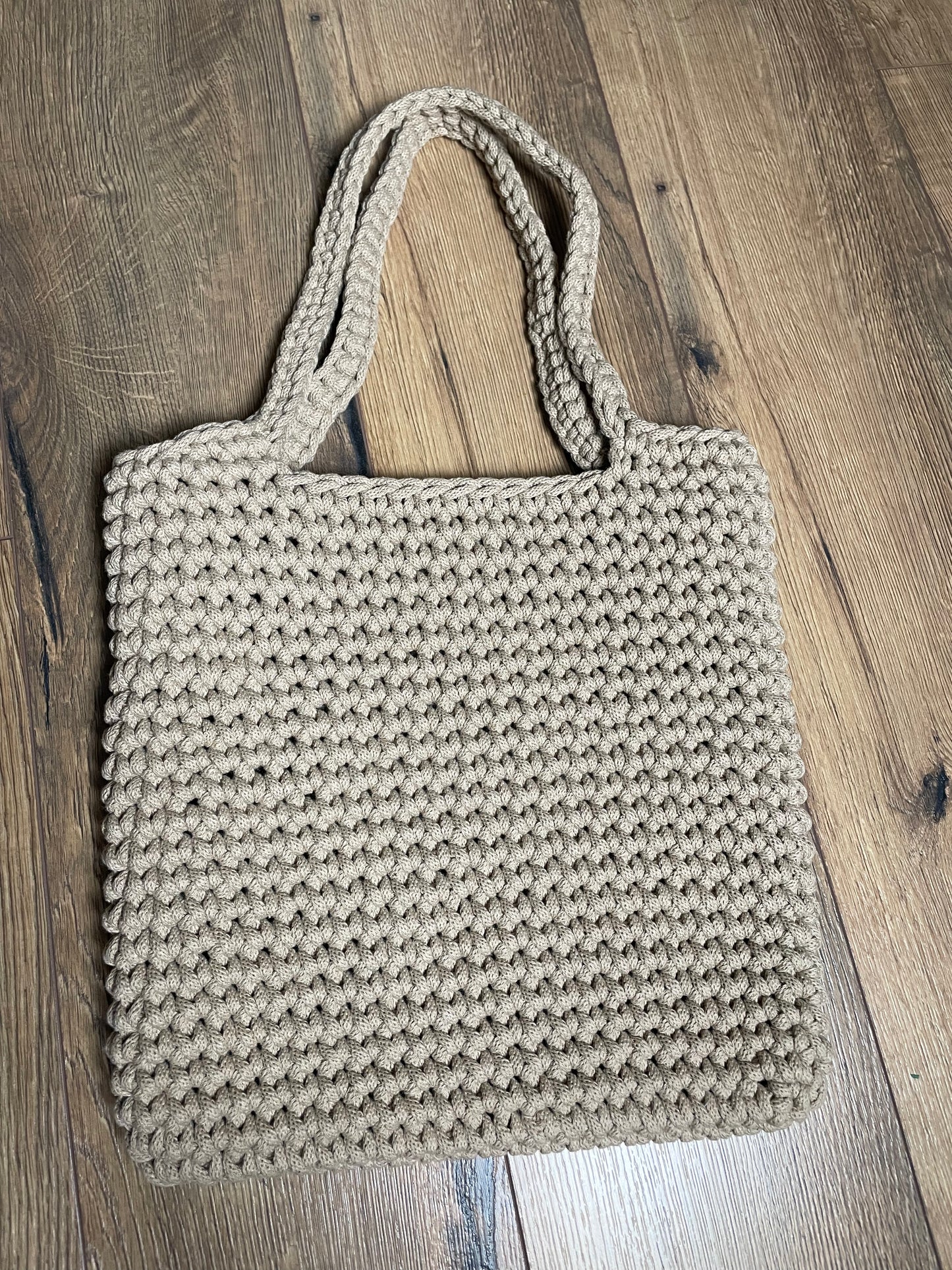 Eine sand-beigefarbene gehäkelte Schultertasche liegt auf Holzlaminatboden. Die Tasche hat ein gleichmäßiges Häkelmuster, zwei lange Henkel und eine rechteckige Form. Das handgefertigte Stück aus recyceltem Baumwollgarn zeigt die charakteristische Struktur der Häkelarbeit mit sichtbaren Maschen.