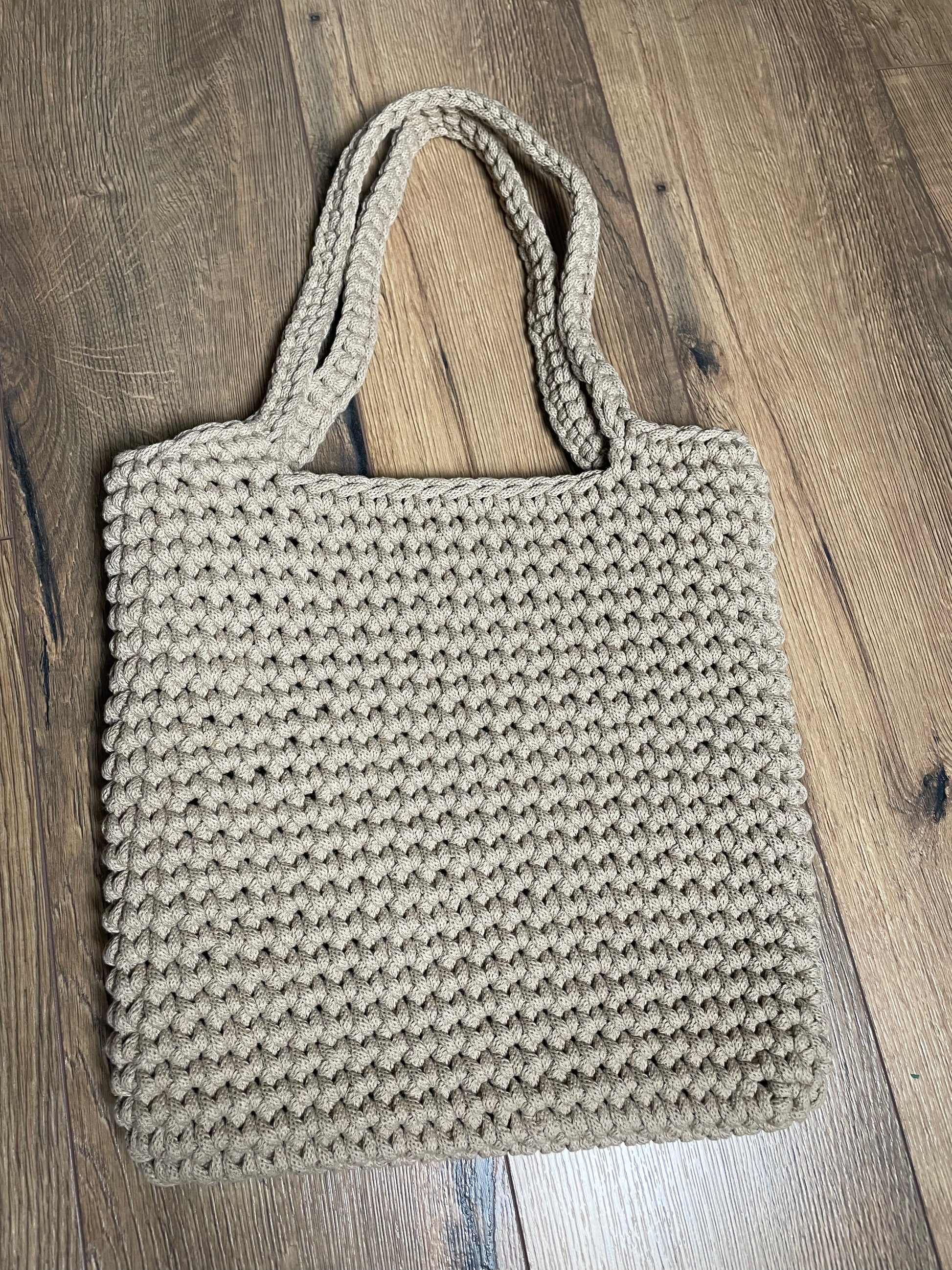 Eine sand-beigefarbene gehäkelte Schultertasche liegt auf Holzlaminatboden. Die Tasche hat ein gleichmäßiges Häkelmuster, zwei lange Henkel und eine rechteckige Form. Das handgefertigte Stück aus recyceltem Baumwollgarn zeigt die charakteristische Struktur der Häkelarbeit mit sichtbaren Maschen.