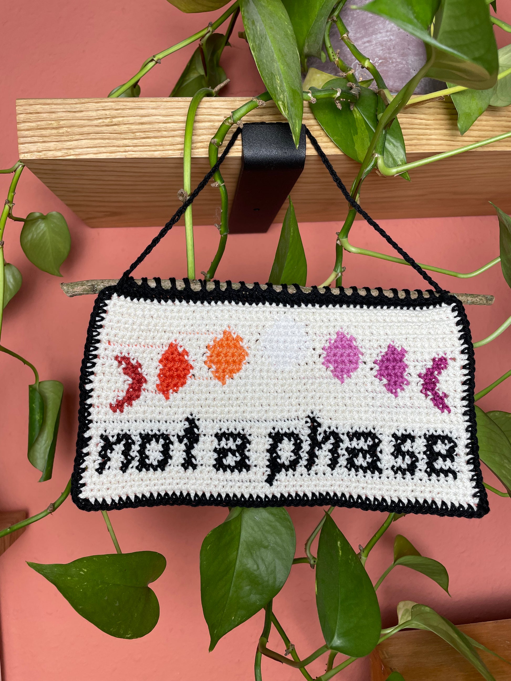 Gehäkeltes Wandbild. Unten in schwarz die Worte "not a phase". Darüber 7 Phasen des Mondes, jeweils in den 7 Farben der lesbischen Pride Flagge. Hintergrund ist cremeweiß. Alles ist umrandet mit schwarz. Am oberen Rand ist ein Stock eingehäkelt. Im Hintergrund eine Efeutute auf einem Holzregal und eine terracottafarbene Wand.