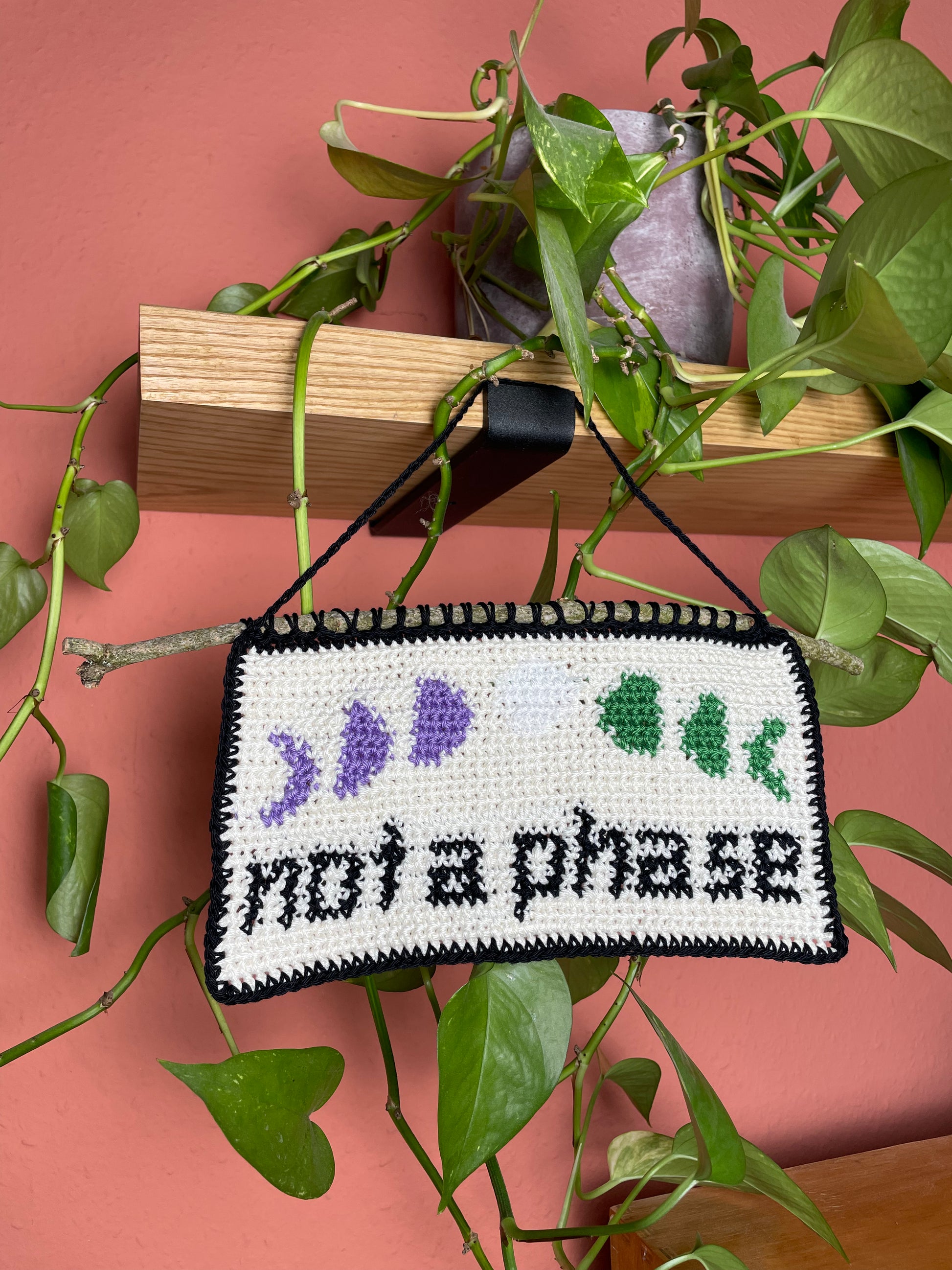 Gehäkeltes Wandbild. Unten in schwarz die Worte "not a phase". Darüber 7 Phasen des Mondes, jeweils in den 3 Farben der Genderqueer/Nonbinary Pride Flagge. Hintergrund ist cremeweiß. Alles ist umrandet mit schwarz. Am oberen Rand ist ein Stock eingehäkelt. Im Hintergrund eine Efeutute auf einem Holzregal und eine terracottafarbene Wand.