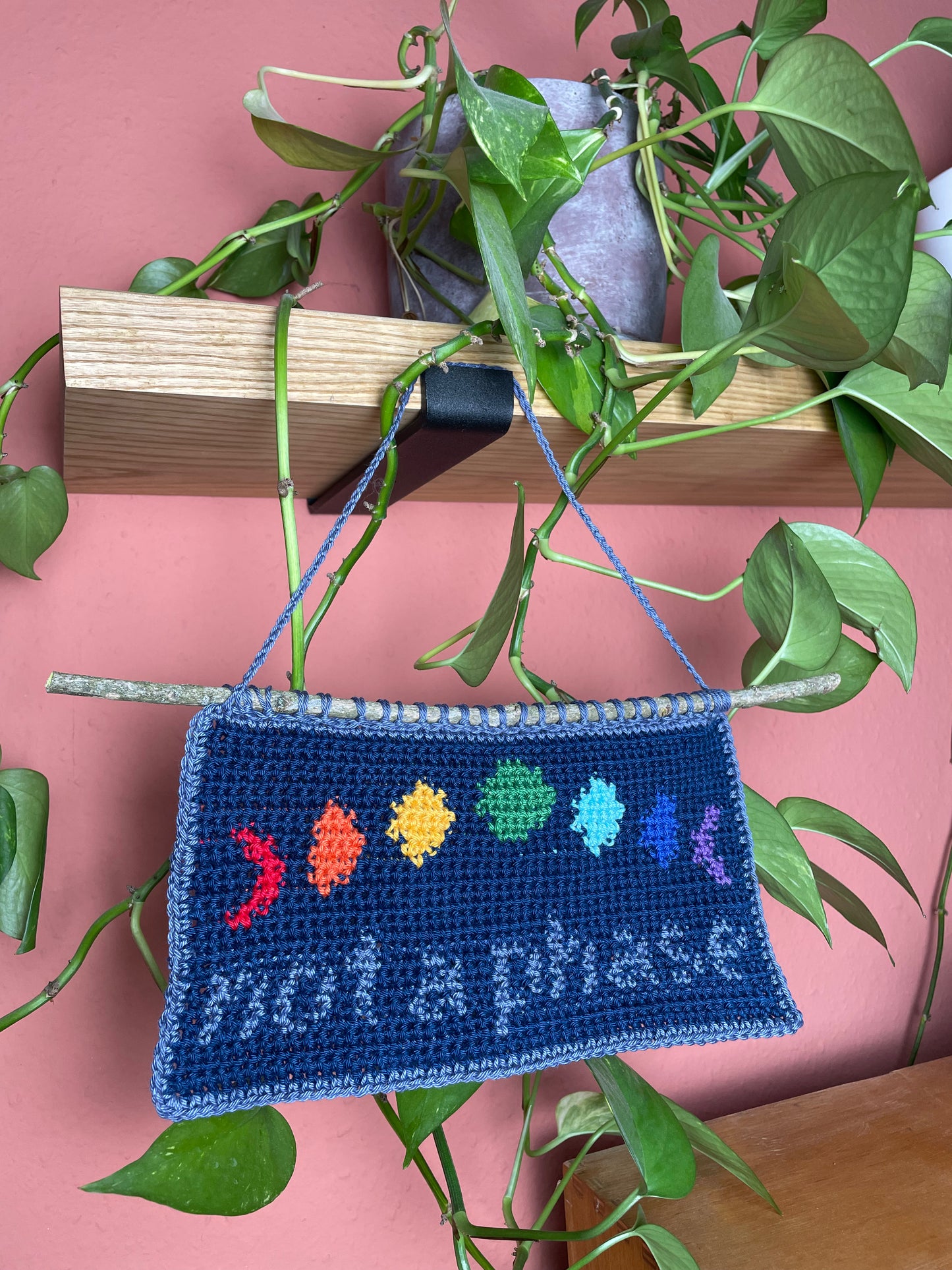 Gehäkeltes Wandbild. Unten in blau-grau die Worte "not a phase". Darüber 7 Phasen des Mondes, jeweils in den 7 Farben des Regenbogens. Hintergrund ist Nachtblau. Alles ist umrandet mit Blaugrau. Am oberen Rand ist ein Stock eingehäkelt. Im Hintergrund eine Efeutute auf einem Holzregal und eine terracottafarbene Wand.