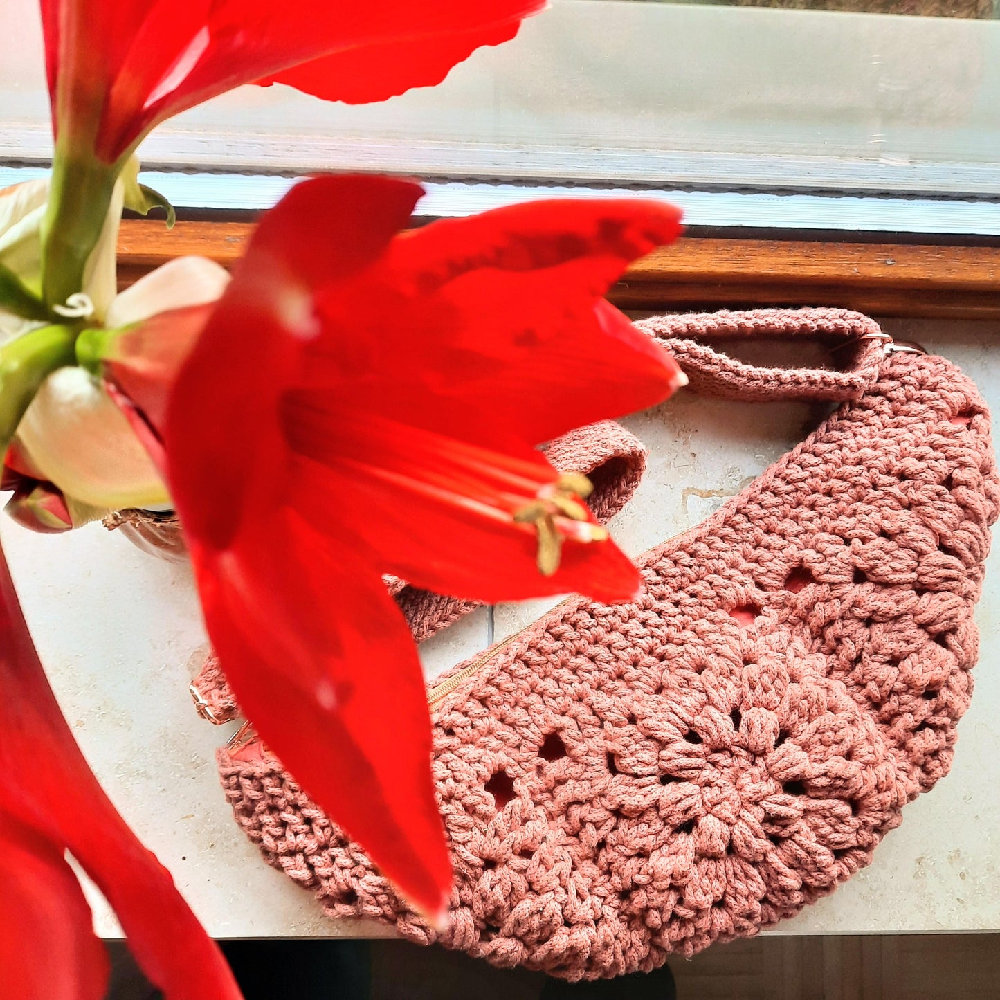 Gehäkelte Tasche bestehend aus vier Granny Squares in Bananenform liegt auf einer Fensterbank, neben und über ihr eine rote blühende Amaryllis.