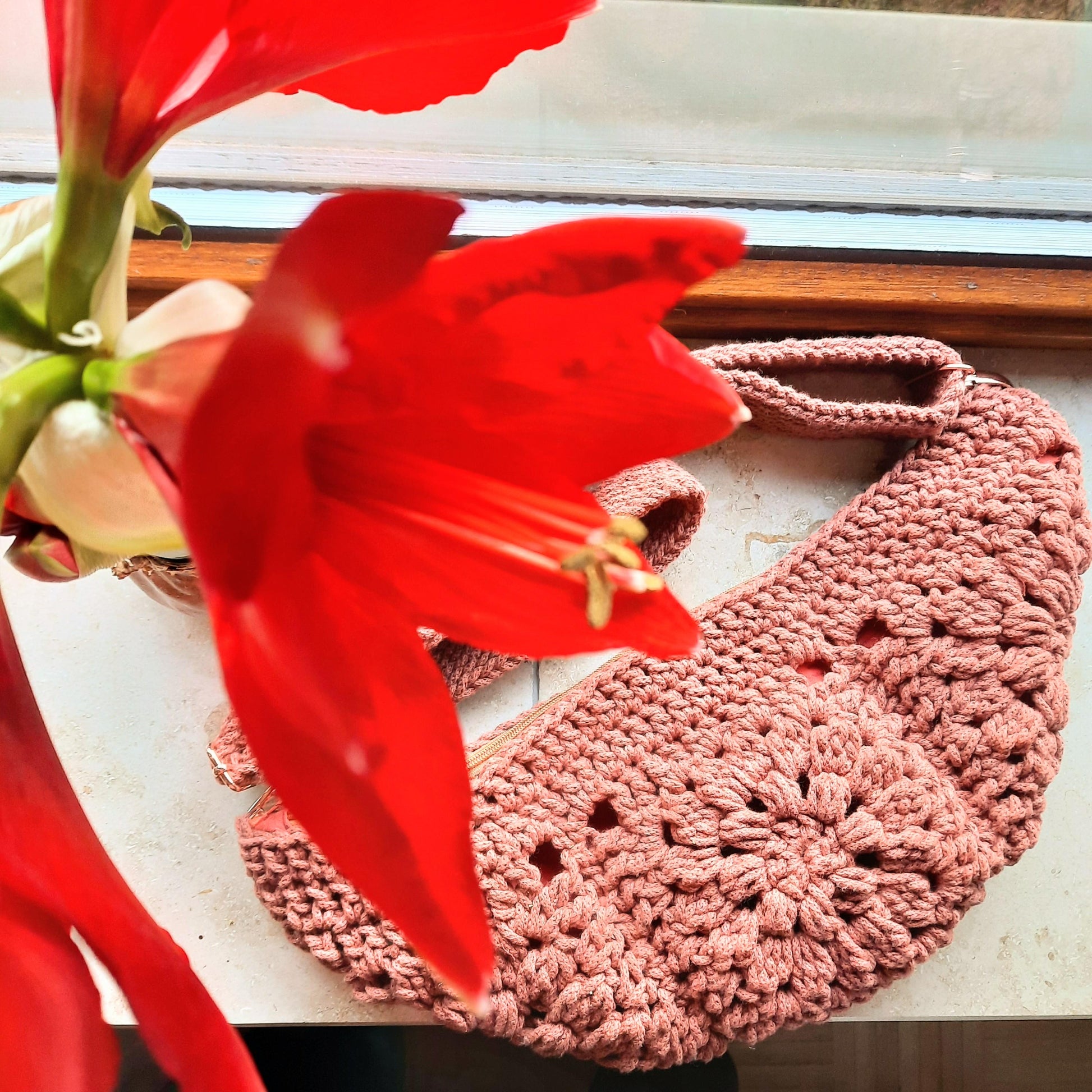 Gehäkelte Tasche bestehend aus vier Granny Squares in Bananenform liegt auf einer Fensterbank, neben und über ihr eine rote blühende Amaryllis.