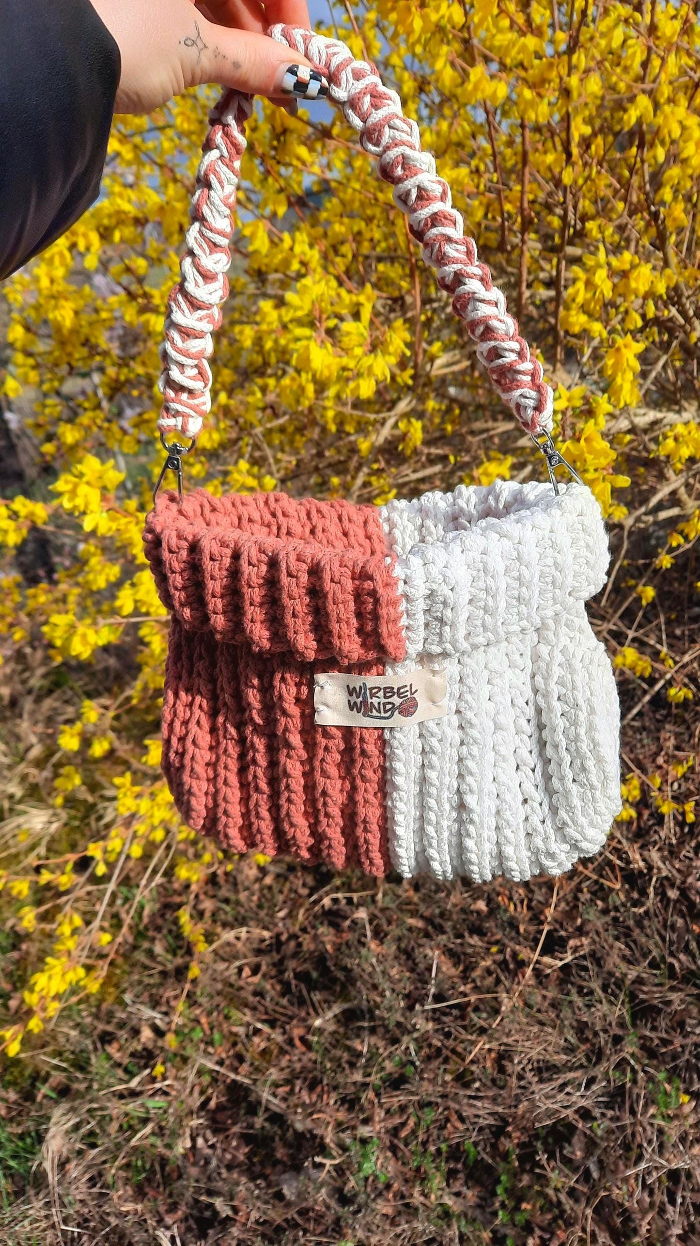 Gabbie Tasche wird am Griff von einer Person ins Bild gehalten, Hand ist oben zu sehen. Sonne scheint. Im Hintergrund gelber Busch, der gerade anfängt zu bühen.