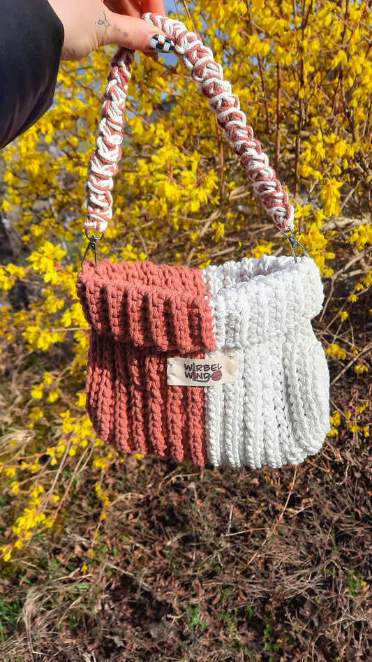 Gabbie Tasche wird am Griff von einer Person ins Bild gehalten, Hand ist oben zu sehen. Sonne scheint. Im Hintergrund gelber Busch, der gerade anfängt zu bühen.