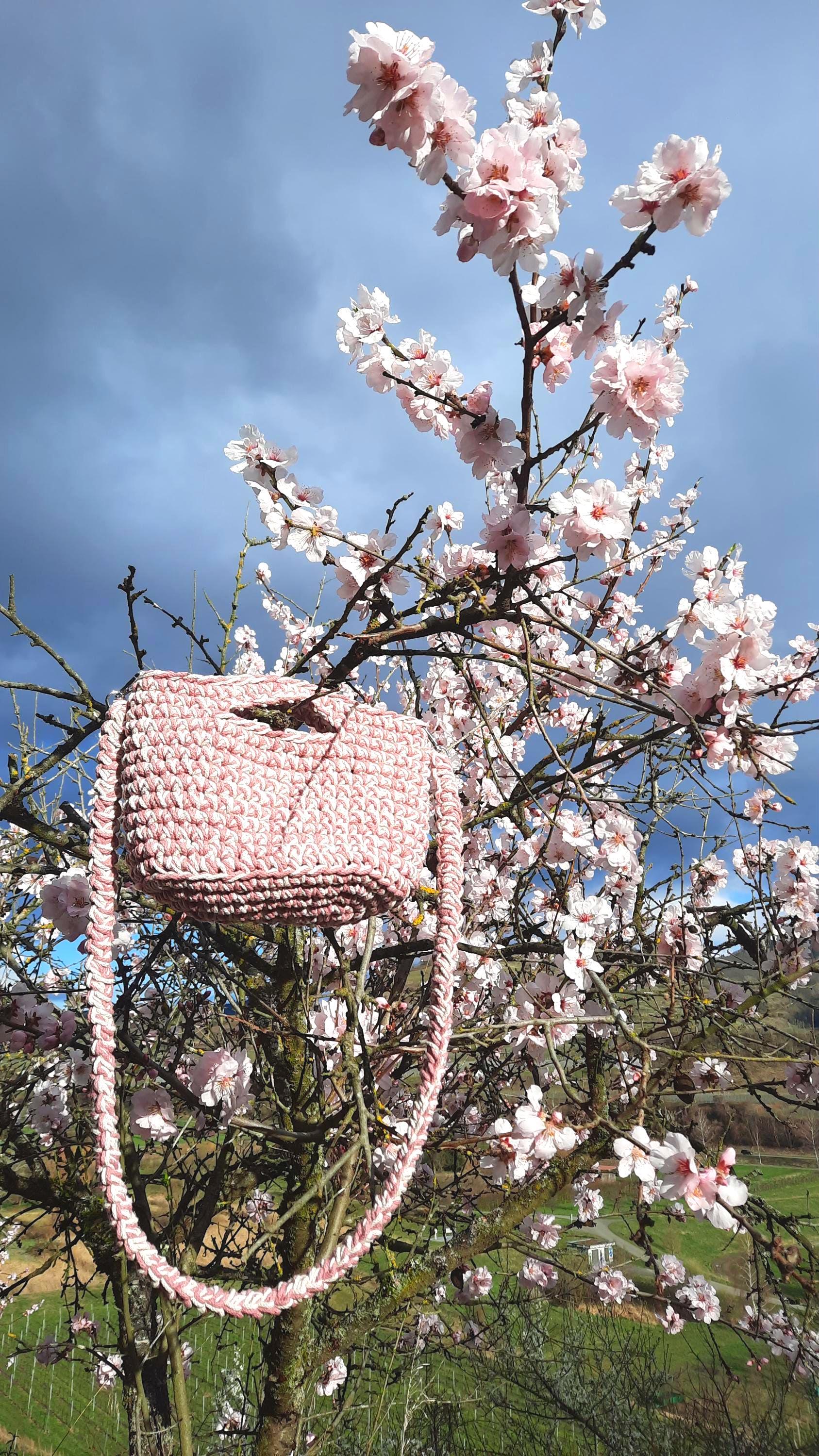 Die rosa-weiße Tasche hängt in einem Kirschbaum mit vielen vollen Blüten in weiß/rosa. Im Hintergrund blauer immel und grüne Wiesen.