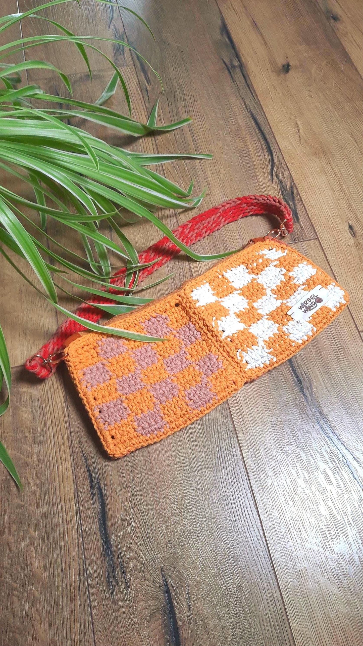 Die Tasche liegt auf Parkett in Holzoptik. Wir sehen die orangene Seite. Der Schultergurt liegt über der Tasche. Am linken Bildrand ragt eine Grünlilie in das Bild.