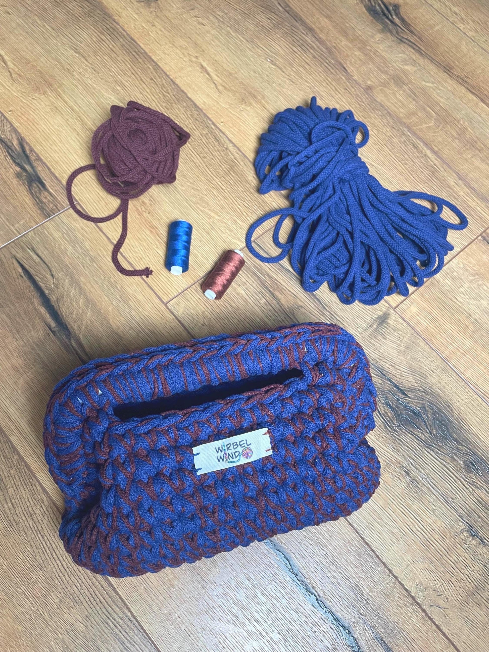 Die Burgunder/Blaue Clutch Tasche liegt auf hölzernem Laminat, neben ihr die beiden Garne, aus der sie gefertigt wurde, sowie zwei farblich passende Garne.