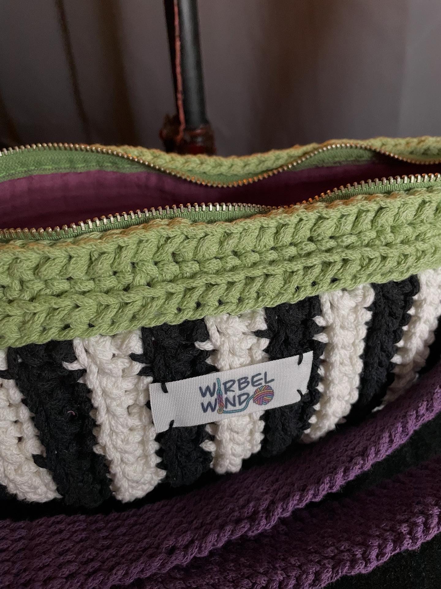 Gehäkelte Tasche in Bananenform in den Farben Schwarz, Weiß, Matcha-Grün und Violet mit verstellbarem Umhängegurt erneut weiter vergrößert. Der Reißverschluss steht offen, wir sehen das violette Innenfutter und die Struktur des Garns. Der Hintergrund ist Anthrazit-Grau, es steht eine schwarz-rote Kerze hinter der Tasche.