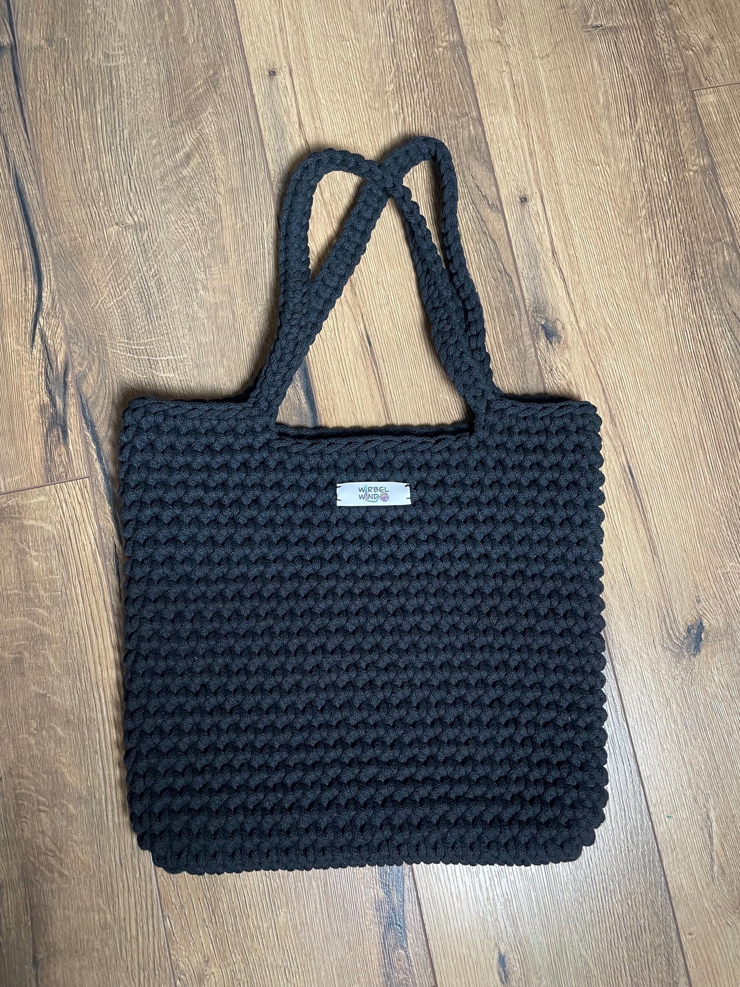 Eine schwarze gehäkelte Schultertasche liegt auf Holzlaminatboden. Die Tasche hat ein gleichmäßiges Häkelmuster, zwei lange Henkel und eine rechteckige Form. Das handgefertigte Stück aus recyceltem Baumwollgarn zeigt die charakteristische Struktur der Häkelarbeit mit sichtbaren Maschen.