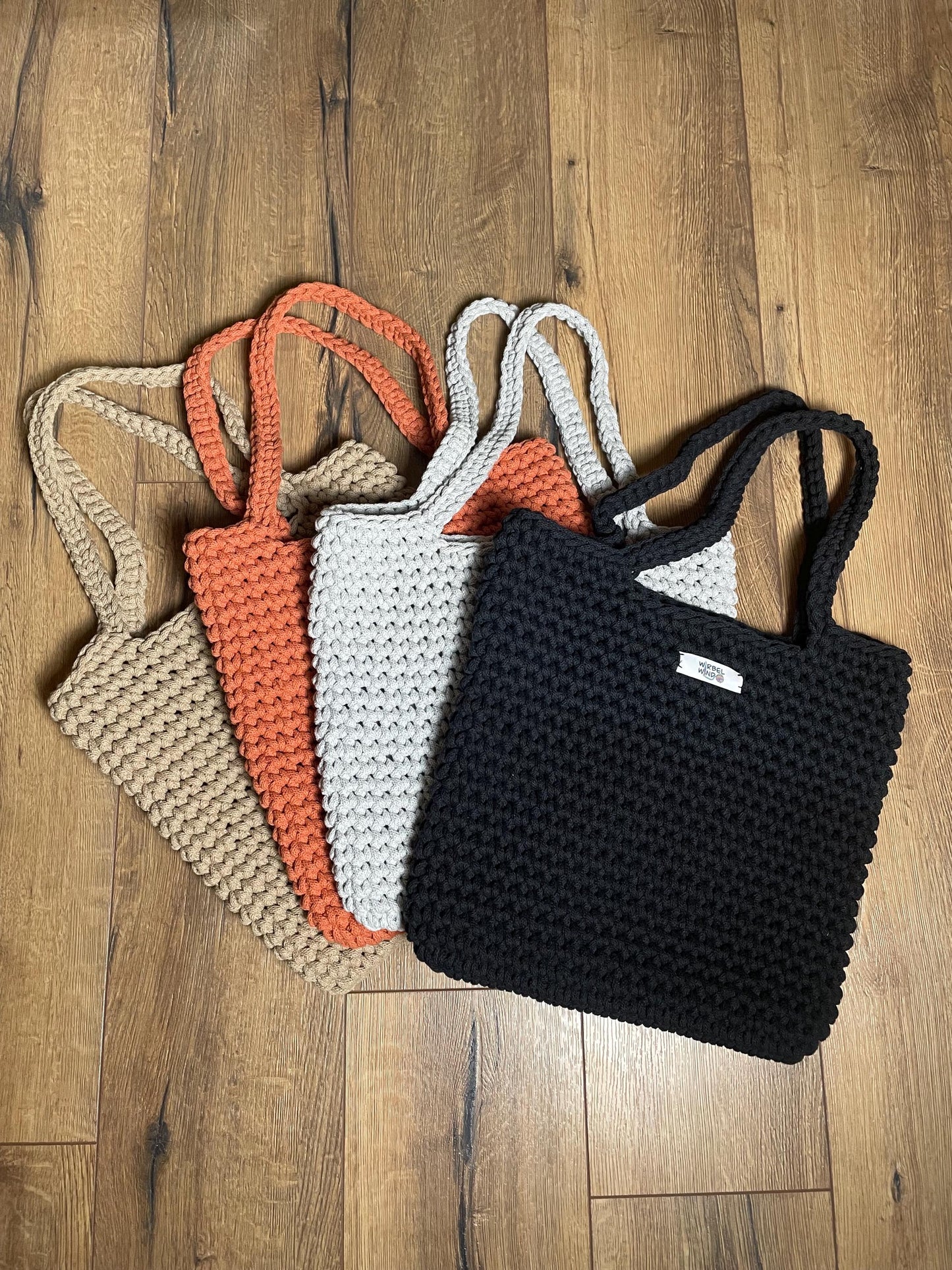 Vier gehäkelte Schultertasche in den Farben Terracotta-Orange, Sand-Beige, Hellgrau und Schwarz liegen aufgefächert auf Holzlaminatboden. Die Taschen haben ein gleichmäßiges Häkelmuster, zwei lange Henkel und eine rechteckige Form. Die handgefertigten Stücke aus recyceltem Baumwollgarn zeigen die charakteristische Struktur der Häkelarbeit mit sichtbaren Maschen.