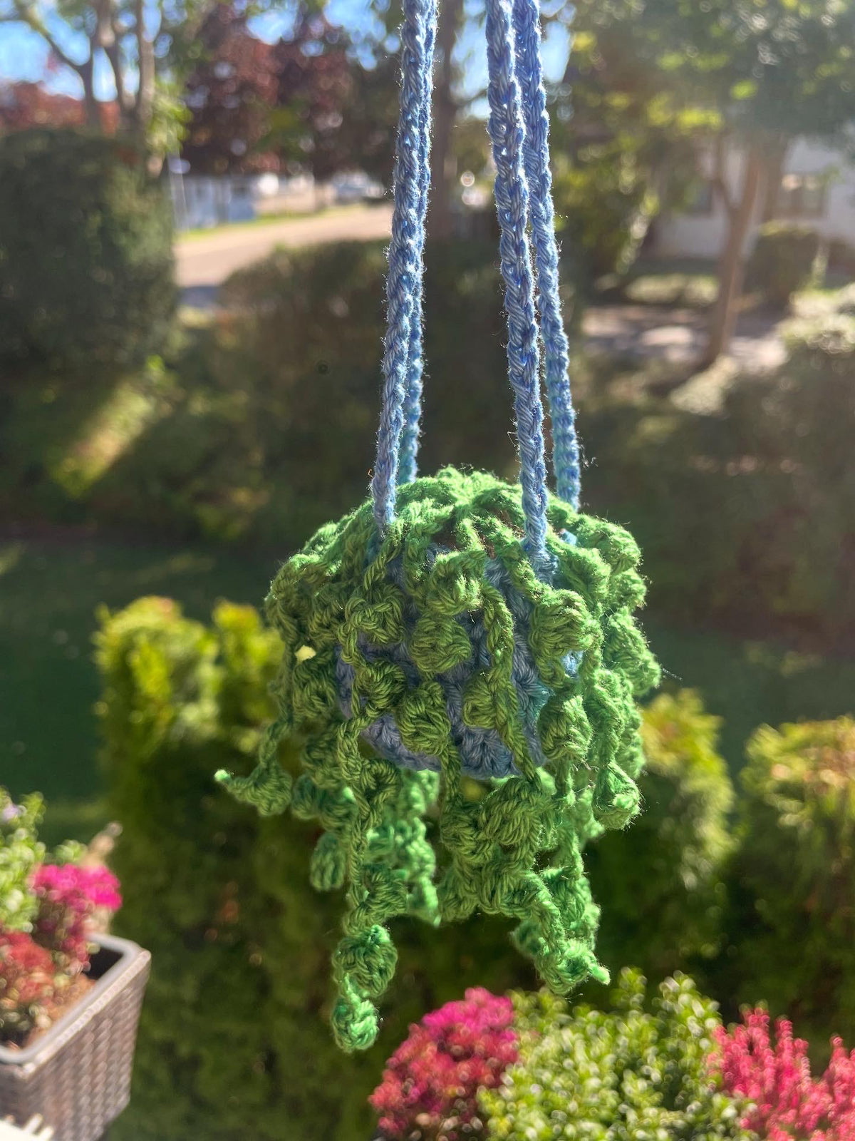 Nahaufnahe einer gehäkelten Erbsenpflanzen in blauem Topf; Hintergrund ist ein Garten, unscharf.